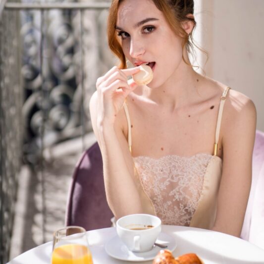 Une jeune femme prend son petit déjeuner au balcon du grand hôtel lyonnais Boscolo, parée d'un caraco et d'un pantalon en soie et dentelle de Calais, réalisés par la Créatrice Aime Couture à Lyon