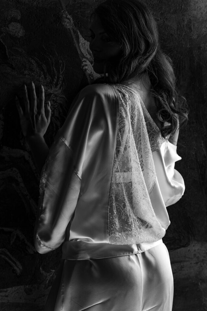 Photo en noir et blanc d'une femme tournée vers un mur dans une chambre du Château de Bagnols, révélant le dos en dentelle de calais du déshabillé Adagio d'Aime Couture
