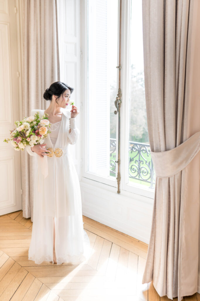 Une jeune femme se tient dans une chambre du Domaine des Halles, devant la fenêtre, en nuisette et déshabillé le jour de son mariage